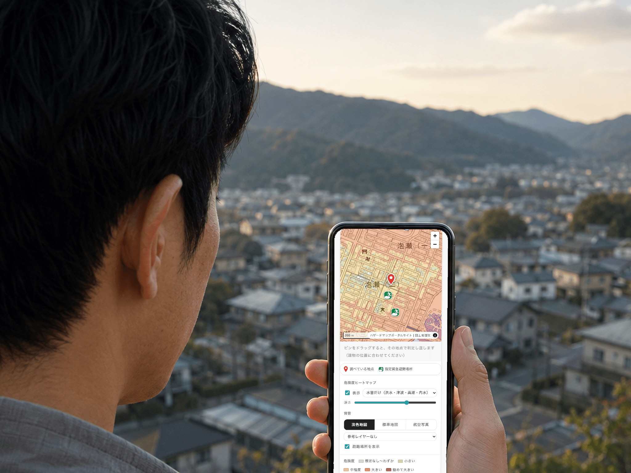 高台から住宅地を見下ろす人が、スマートフォンでソコ調の危険度ヒートマップを開いている。地図には調べている地点のピンと、近くの指定緊急避難場所が表示されている