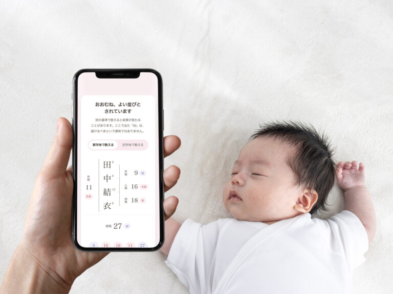 眠っている赤ちゃんの横で、スマートフォンに表示した「なまえのたね」の判定結果を見ている。画面には五格を縦書きで並べた図が出ている